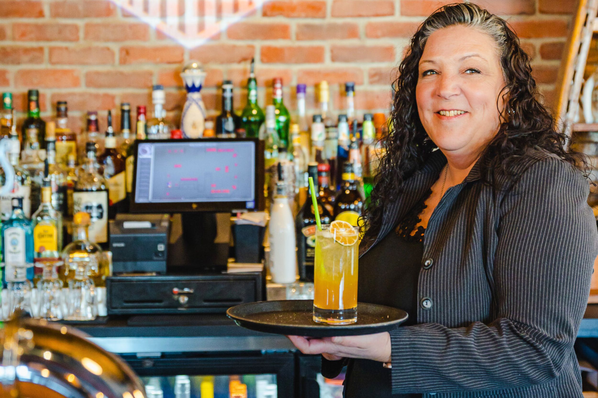 A white dark haired woman with curly hair is wearing a black shirt and a white and black striped overshirt. She is standing in front of a bar while holding a try with a drink on it.